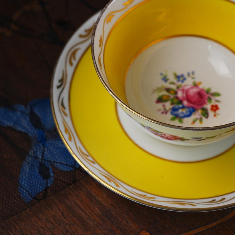 Op de afbeelding: Een porseleinen theekop en schotel set met een felgele rand en gouden afwerking. De binnenkant van de kop en het midden van de schotel zijn wit, versierd met een kleurrijk bloemenboeket van roze rozen en kleine blauwe en rode bloemetjes.