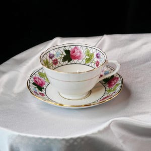 Op de afbeelding: Witte theekop en schotel set met een rozenpatroon. De theekop en schotel hebben gouden randen en een zwart traliewerk patroon. De theekop heeft een oor en staat op een witte satijnen ondergrond.