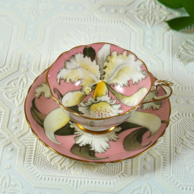 May include: A pink teacup and saucer set with a floral design. The cup and saucer are adorned with a large, white and yellow orchid-like flower. The set has gold trim and is set on a white patterned surface.