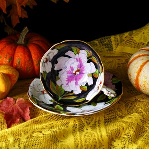 May include: A floral teacup and saucer set with a black background and gold trim. The teacup and saucer feature a large white and pink peony design with green leaves. The set is placed on a yellow lace tablecloth, with pumpkins in the background.