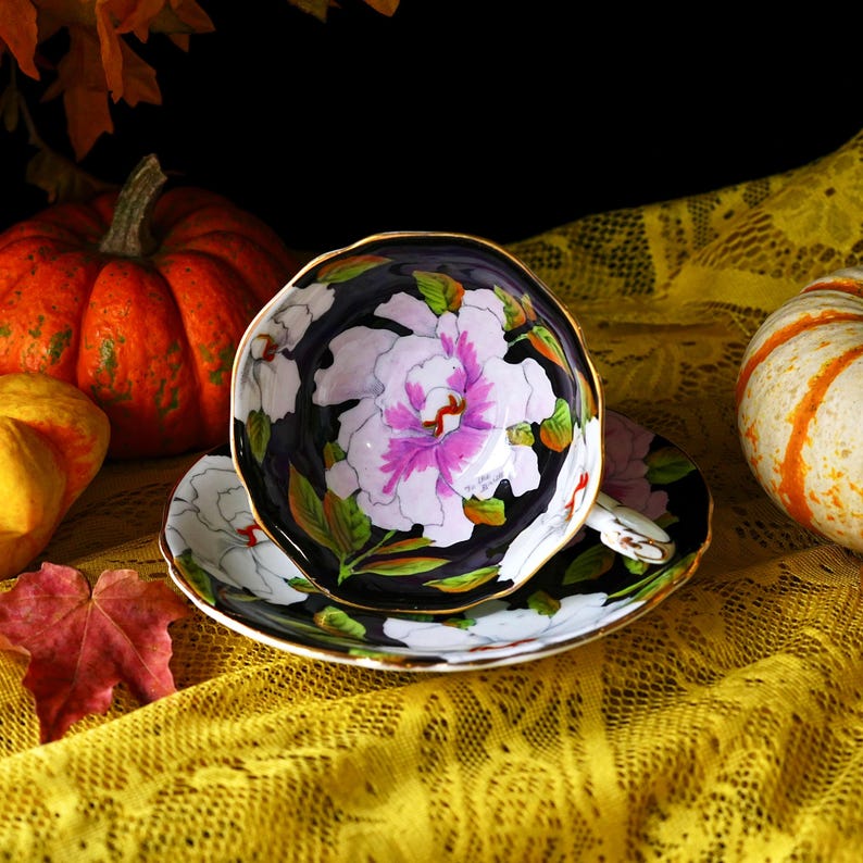 May include: A floral teacup and saucer set with a black background and gold trim. The teacup and saucer feature a large white and pink peony design with green leaves. The set is placed on a yellow lace tablecloth, with pumpkins in the background.