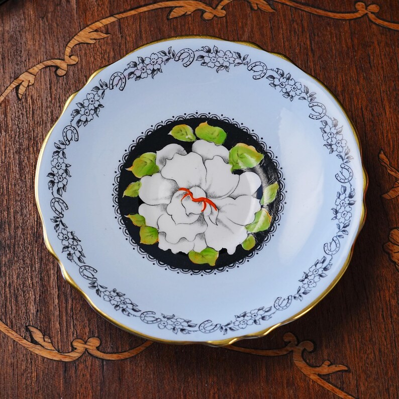 May include: A round porcelain saucer with a scalloped gold-trimmed edge. The center features a large white flower with green leaves on a black circular background. The outer rim is light blue with a black floral and horseshoe pattern border.