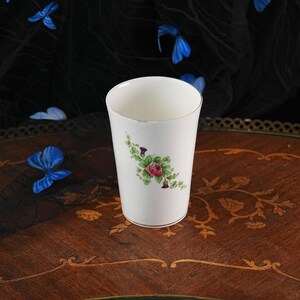 Puede incluir: Un vaso de cer&aacute;mica blanca con un delicado motivo floral de una rosa rosa rodeada de hojas verdes y peque&ntilde;os capullos morados similares a cardos. El recipiente cil&iacute;ndrico tiene un acabado liso y un borde met&aacute;lico fino en los extremos.