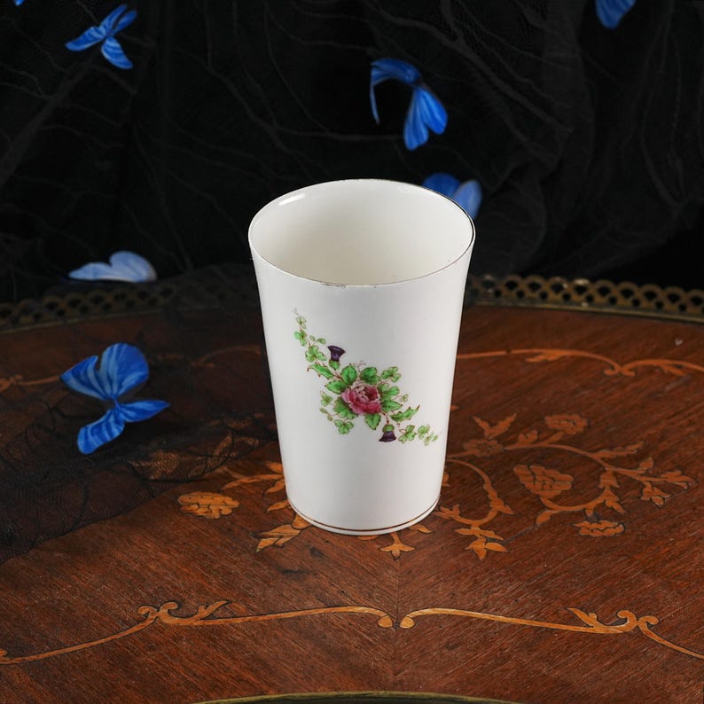 Puede incluir: Un vaso de cer&aacute;mica blanca con un delicado motivo floral de una rosa rosa rodeada de hojas verdes y peque&ntilde;os capullos morados similares a cardos. El recipiente cil&iacute;ndrico tiene un acabado liso y un borde met&aacute;lico fino en los extremos.