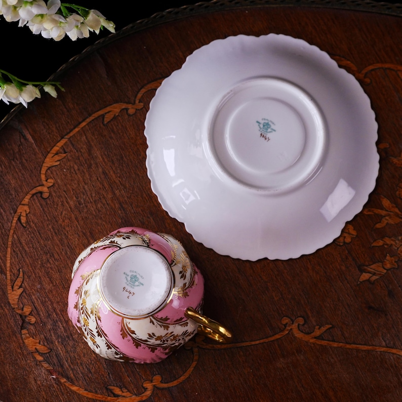 May include: A vintage teacup and saucer set. The teacup features a pink and gold floral design with a gold handle. The saucer is white with a scalloped edge. The set is placed on a dark wooden surface.