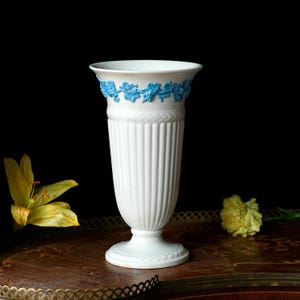 May include: A white ceramic vase with a fluted body and a blue floral design around the rim. The vase has a pedestal base and is displayed on a brown surface with yellow flowers.