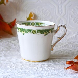 May include: White teacup with a green floral band and gold trim. The handle is also gold. The cup has a classic design, suitable for tea or coffee. The cup is on a white lace tablecloth.