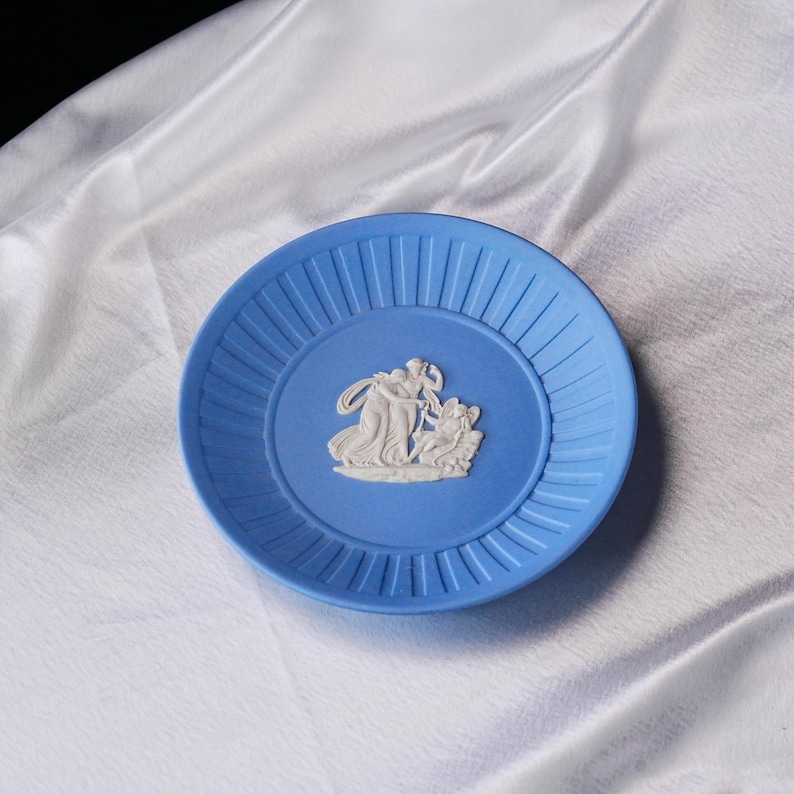 May include: A round, light blue ceramic plate featuring a white, raised relief design in the center depicting two figures and a winged cherub. The plate has a wide, flat rim with vertical, evenly spaced grooves encircling the central scene.