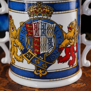 May include: A white ceramic mug featuring a colourful royal coat of arms with a central shield, a crown, and two lions on either side. The design includes a blue garter with the text HONI SOIT QUI MAL Y PENSE and ornate white handles.