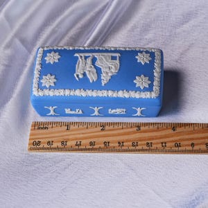 May include: A blue rectangular trinket box with white floral and classical relief designs. The box measures approximately 3 inches (7.6 cm) in length. The lid features a central relief scene and floral accents.