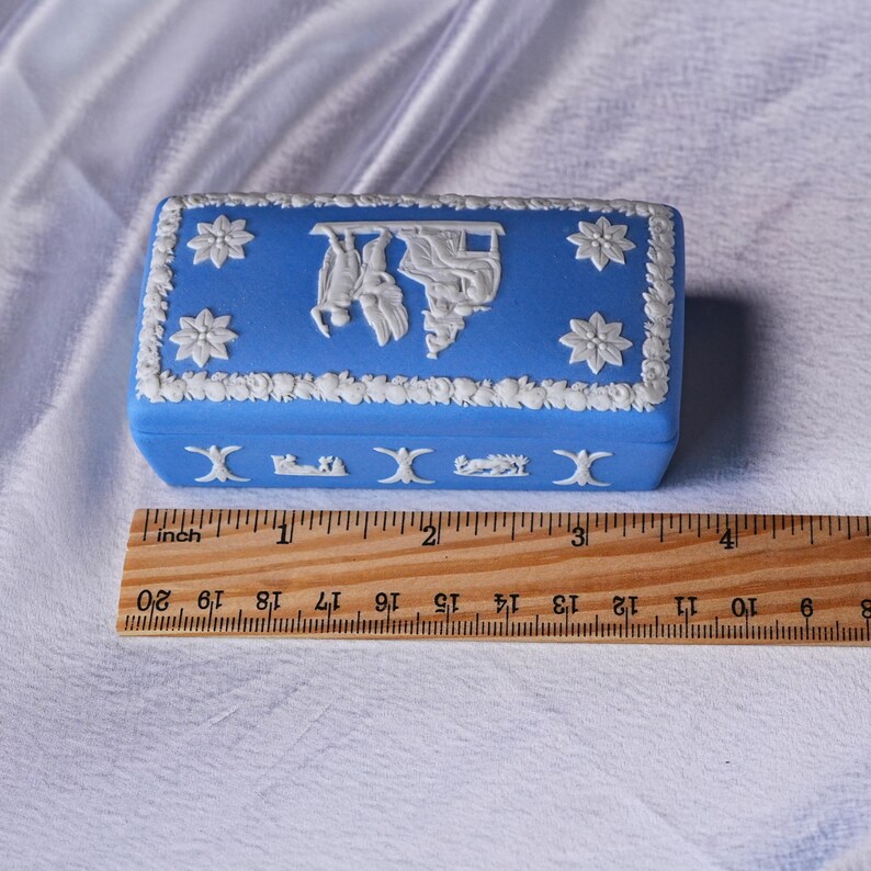 May include: A blue rectangular trinket box with white floral and classical relief designs. The box measures approximately 3 inches (7.6 cm) in length. The lid features a central relief scene and floral accents.