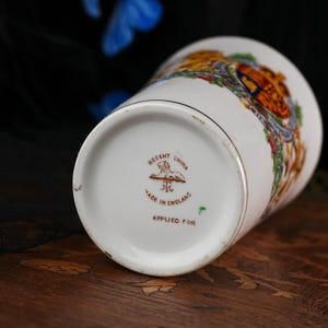 Puede incluir: Una taza de cer&aacute;mica blanca con un escudo colorido en el lateral. La base muestra el texto 'REGENT CHINA', un emblema de le&oacute;n, 'MADE IN ENGLAND' y 'APPLIED FOR' en tinta marr&oacute;n. La taza tiene una forma cil&iacute;ndrica con un borde dorado.