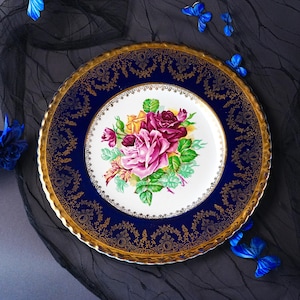 May include: A round ceramic plate featuring a white center decorated with a cluster of pink, purple, and yellow roses with green leaves. The wide border is deep blue with an intricate gold filigree pattern and a scalloped, gilded edge.