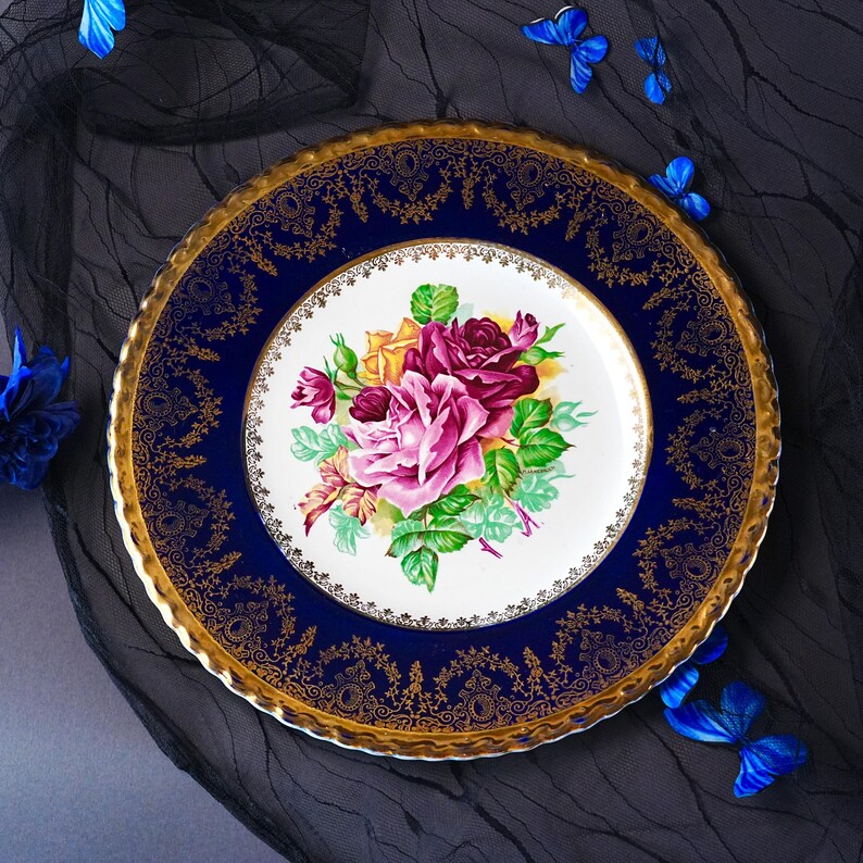 May include: A round ceramic plate featuring a white center decorated with a cluster of pink, purple, and yellow roses with green leaves. The wide border is deep blue with an intricate gold filigree pattern and a scalloped, gilded edge.