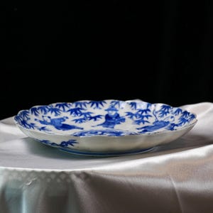 Puede incluir: Un plato de cer&aacute;mica blanco con borde festoneado, con una ilustraci&oacute;n azul y blanca de figuras y bamb&uacute;. El plato tiene un pie ligeramente elevado y est&aacute; sobre una tela blanca.
