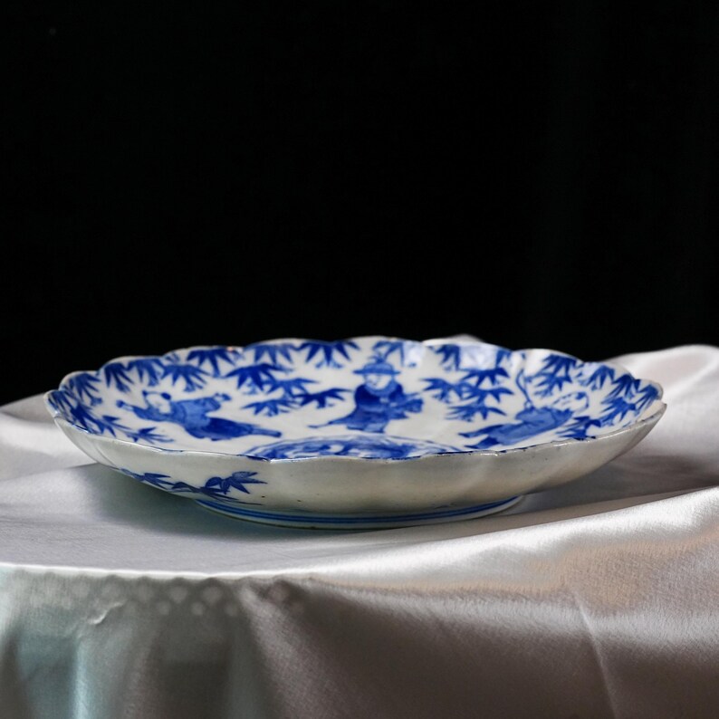 Puede incluir: Un plato de cer&aacute;mica blanco con borde festoneado, con una ilustraci&oacute;n azul y blanca de figuras y bamb&uacute;. El plato tiene un pie ligeramente elevado y est&aacute; sobre una tela blanca.