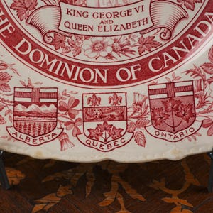 May include: A cream-coloured ceramic plate featuring red transferware designs. The text reads KING GEORGE VI AND QUEEN ELIZABETH THE DOMINION OF CANADA. Below are the coats of arms for Alberta, Quebec, and Ontario with their names printed below.