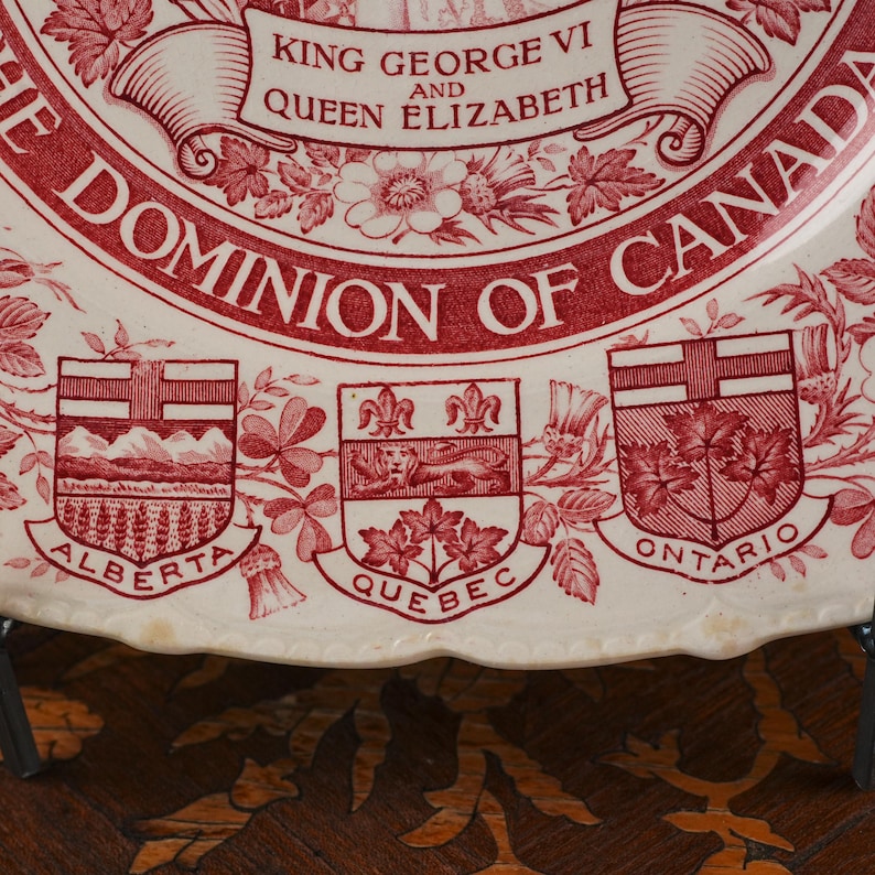 May include: A cream-coloured ceramic plate featuring red transferware designs. The text reads KING GEORGE VI AND QUEEN ELIZABETH THE DOMINION OF CANADA. Below are the coats of arms for Alberta, Quebec, and Ontario with their names printed below.