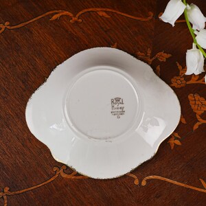 May include: The underside of a white ceramic saucer with a scalloped edge and gold trim. The center features a black stamp reading Royal Harvey, Staffordshire, England. The saucer has a circular base and a smooth, glossy finish on the white surface.