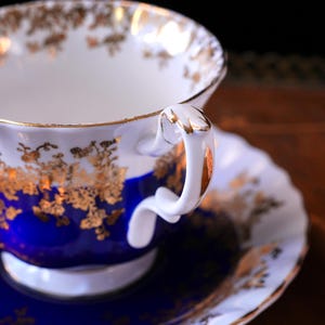 May include: A detailed shot of a porcelain teacup and saucer. The cup and saucer are white with gold floral designs and gold edging. The cup's base is a deep blue, and the handle is white with gold detailing. The saucer is also blue.