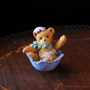 May include: A charming figurine of a teddy bear emerging from a blue and white eggshell. The bear wears a pink hat and a blue collar. The eggshell has a scalloped edge and is decorated with small pink dots. The bear is holding up one paw.