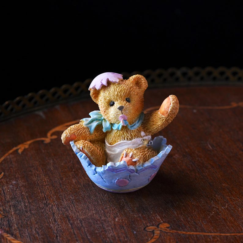 May include: A charming figurine of a teddy bear emerging from a blue and white eggshell. The bear wears a pink hat and a blue collar. The eggshell has a scalloped edge and is decorated with small pink dots. The bear is holding up one paw.
