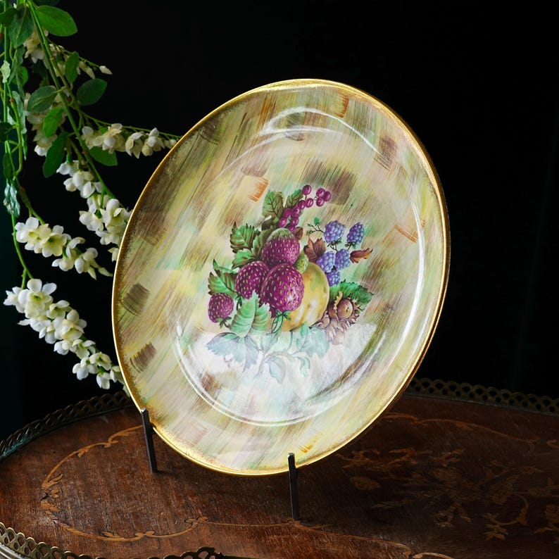 May include: An oval-shaped decorative plate with a gold rim, featuring a hand-painted still life of purple berries, a yellow fruit, and green leaves. The plate has a textured, light green and yellow background.