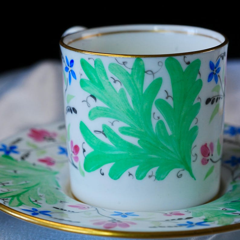 May include: A white porcelain teacup and saucer set featuring a hand-painted design of large green leaves, small blue flowers, and pink floral accents. The rim of both the cup and the saucer is finished with a thin, metallic gold painted edge.