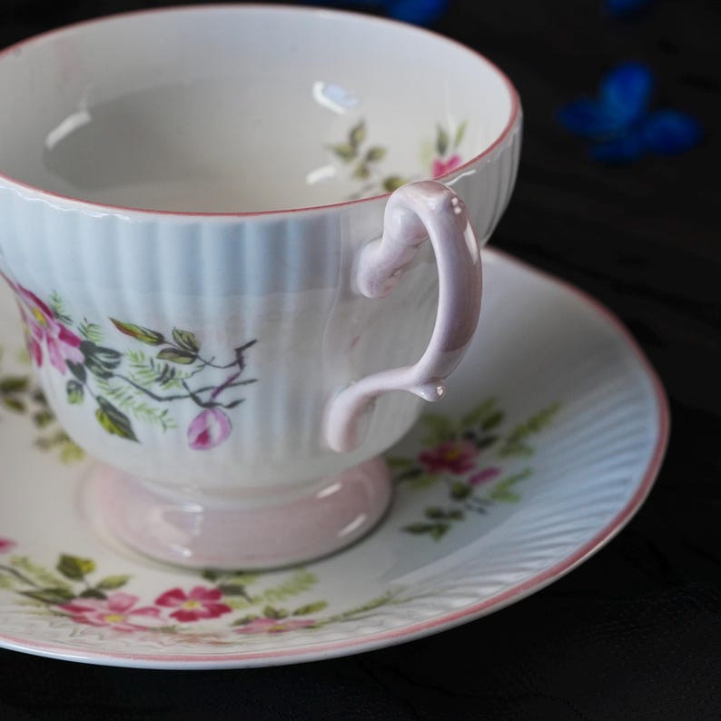 May include: A white porcelain teacup and saucer set featuring a delicate floral pattern with pink and green botanical illustrations. The cup has a fluted exterior, a light pink handle, and a matching pink rim on both the cup and the saucer.