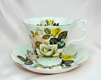 May include: A white teacup and saucer set with a floral pattern. The pattern features pink, yellow, and green roses with brown leaves. The cup has a gold rim and a handle.