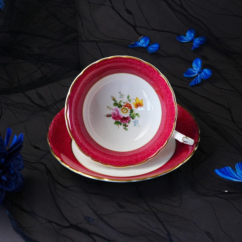 K&ouml;nnte beinhalten: Ein Porzellan-Tassenset mit Untertasse, das einen leuchtend magentafarbenen Rand mit strukturiertem Muster und Goldverzierung aufweist. Das Innere der Tasse und die Mitte der Untertasse sind wei&szlig; und mit einem Blumenstrau&szlig; in Rosa, Gelb und Gr&uuml;n verziert.