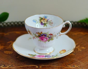 May include: A white teacup and saucer set with a floral design. The teacup has a gold rim and handle, and the saucer has a scalloped edge. The floral pattern includes pink, purple, yellow, and blue flowers.