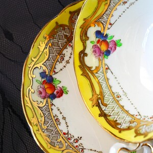 Peut inclure: Un ensemble tasse et soucoupe en porcelaine blanche avec une bordure jaune vif, des volutes dor&eacute;es complexes et un motif floral peint &agrave; la main avec des fleurs roses, orange et bleues. Le design inclut un fin motif en losanges noirs.