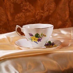 K&ouml;nnte beinhalten: Ein wei&szlig;es Tee-Tassen- und Untertassen-Set mit einem Blumendesign mit gelben, lila und gr&uuml;nen Blumen. Die Tasse und die Untertasse haben einen goldenen Rand. Das Set steht auf einer Satin-Oberfl&auml;che.