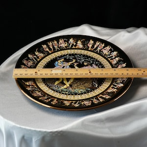 May include: A black decorative plate with a gold rim, featuring a detailed illustration of figures in classical attire. The plate measures approximately 10 inches in diameter. The artwork depicts a scene with archers and other figures.