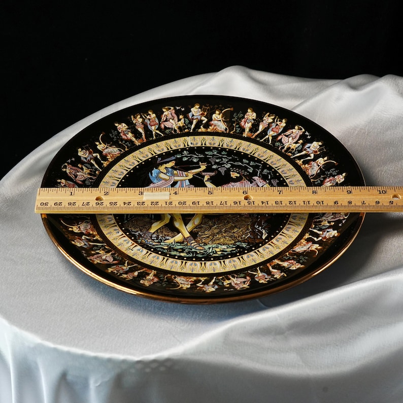 May include: A black decorative plate with a gold rim, featuring a detailed illustration of figures in classical attire. The plate measures approximately 10 inches in diameter. The artwork depicts a scene with archers and other figures.