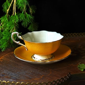 May include: A yellow teacup and saucer set with gold detailing. The teacup has a white interior and a decorative handle. The saucer is round and matches the cup's colour. The set is displayed on a wooden tray, evoking a sense of classic elegance and afternoon tea.