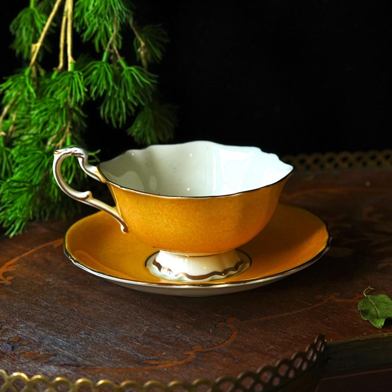 May include: A yellow teacup and saucer set with gold detailing. The teacup has a white interior and a decorative handle. The saucer is round and matches the cup's colour. The set is displayed on a wooden tray, evoking a sense of classic elegance and afternoon tea.