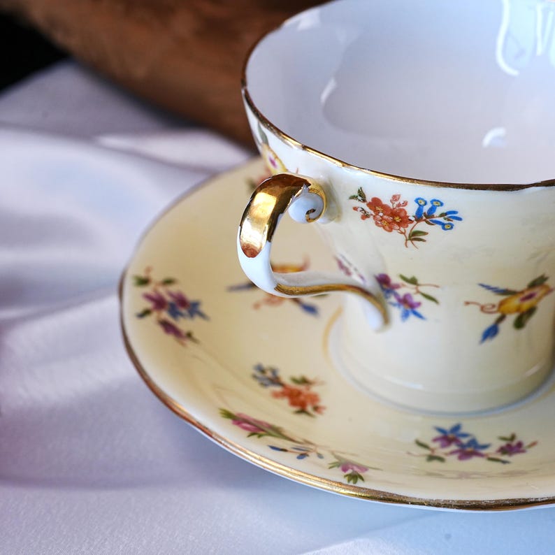 Op de afbeelding: Een vintage theekop en schotel set. De kop en schotel zijn cr&egrave;mekleurig met delicate bloempatronen in rood, blauw en paars. De handgreep en rand van de kop zijn goudkleurig afgewerkt. De set staat op een witte ondergrond.