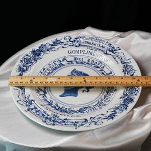 May include: White and blue commemorative plate with a portrait of a woman, possibly Queen Victoria, and the text "Jubilee Year Compliments of Dei Gratia". The plate is approximately 12 inches in diameter.