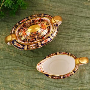 May include: An oval-shaped, covered serving dish and a gravy boat. The porcelain pieces feature a gold rim and handles, with a detailed floral and geometric pattern in red, blue, and gold. The set is placed on a green cloth.