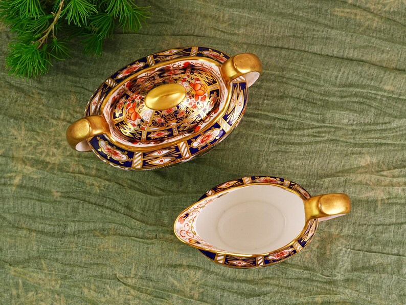 May include: An oval-shaped, covered serving dish and a gravy boat. The porcelain pieces feature a gold rim and handles, with a detailed floral and geometric pattern in red, blue, and gold. The set is placed on a green cloth.