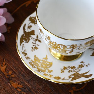Op de afbeelding: Witte theekop en schotel set met gouden bloemen- en vogelontwerpen. De theekop heeft een gouden voet en rand, en de schotel heeft bijpassende gouden details. Een klassieke theeset.