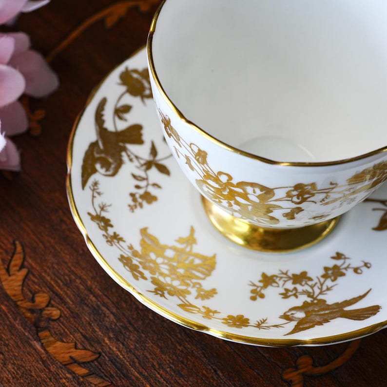 Op de afbeelding: Witte theekop en schotel set met gouden bloemen- en vogelontwerpen. De theekop heeft een gouden voet en rand, en de schotel heeft bijpassende gouden details. Een klassieke theeset.