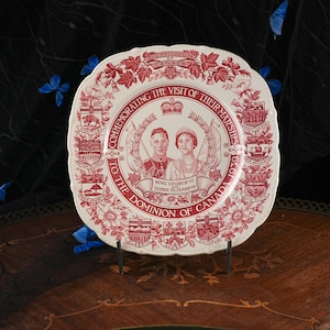 May include: A cream-coloured ceramic plate with a red transfer print commemorating the 1939 visit of King George VI and Queen Elizabeth to Canada. The plate features portraits of the monarchs, the text Dominion of Canada, and provincial crests.