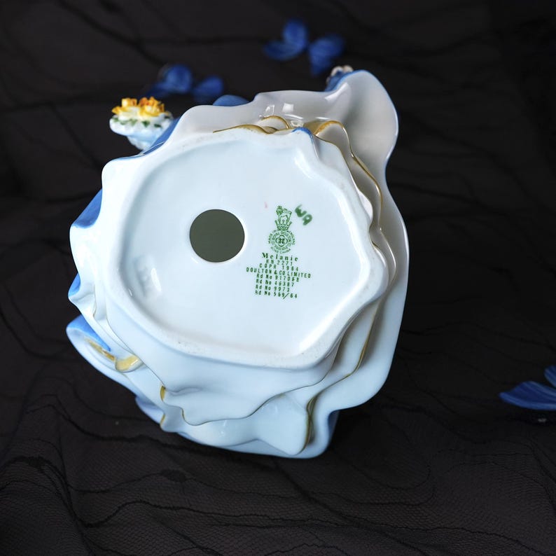 May include: A porcelain figurine base showing the underside with green printed text reading Melanie, HN 2271, Copr 1964, Doulton & Co Limited, and various registration numbers. The base is white with a circular hole and blue ruffled edges with gold trim.