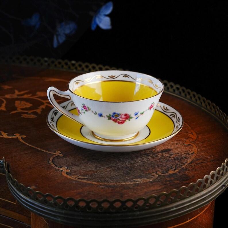 Op de afbeelding: Een porseleinen theekop en schotel set met een felgele binnenkant en middenstuk. De buitenkant van de kop en de rand van de schotel zijn versierd met een delicate bloemenslinger van roze rozen en blauwe vergeet-mij-nietjes met gouden rand.