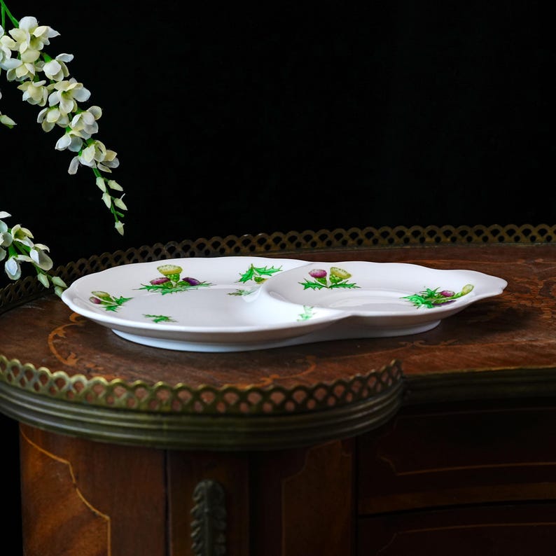 Puede incluir: Plato de servir blanco dividido con borde festoneado. El plato est&aacute; decorado con hojas verdes de acebo y flores de cardo moradas y amarillas. El plato est&aacute; sobre una superficie de madera oscura.