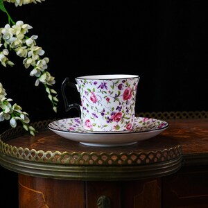May include: A floral teacup and saucer set featuring pink roses, purple accents, and green foliage on a white background. The cup has a black handle and trim. The set rests on a wooden table with a decorative metal edge.