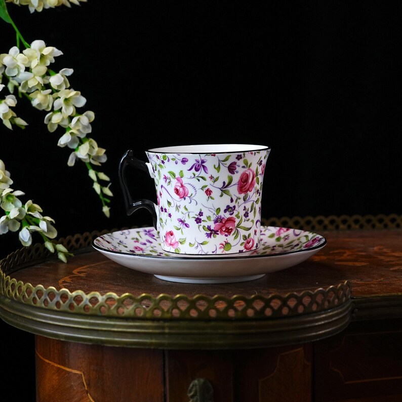 May include: A floral teacup and saucer set featuring pink roses, purple accents, and green foliage on a white background. The cup has a black handle and trim. The set rests on a wooden table with a decorative metal edge.
